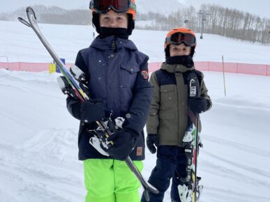 2 young kids in snow outfits holding their skiis at the base of a mountain